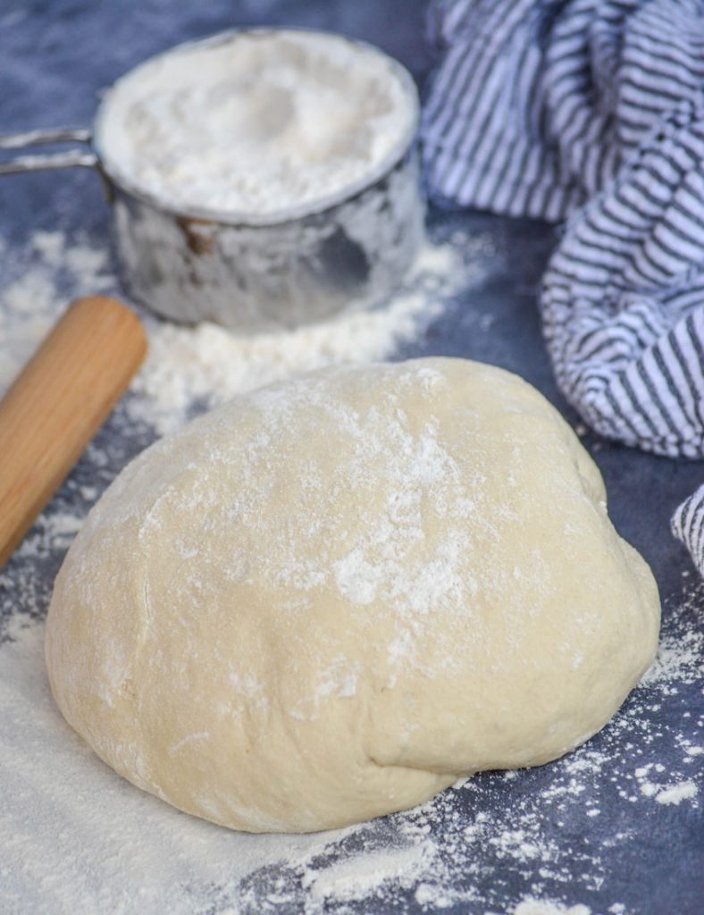 a ball of floured easy homemade pizza dough is shown on a gray back ground with a wooden rolling pin, excess flour, and a striped dish cloth in the back ground
