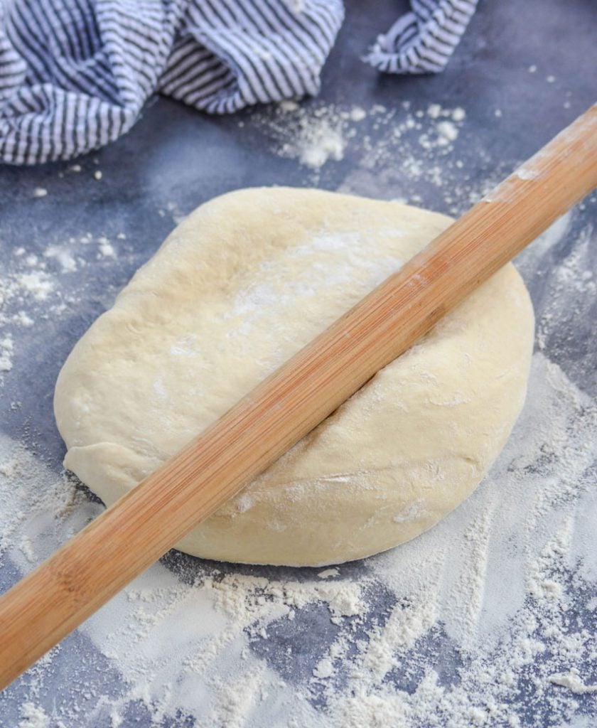 floured pizza dough ball being rolled out by a wooden rolling pin on a gray back ground