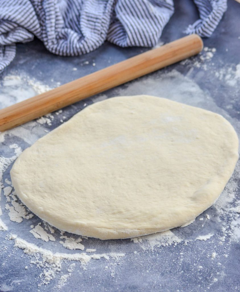 semi rolled out pizza dough on a floured gray surface with a wooden rolling pin and striped cloth napkin in the background