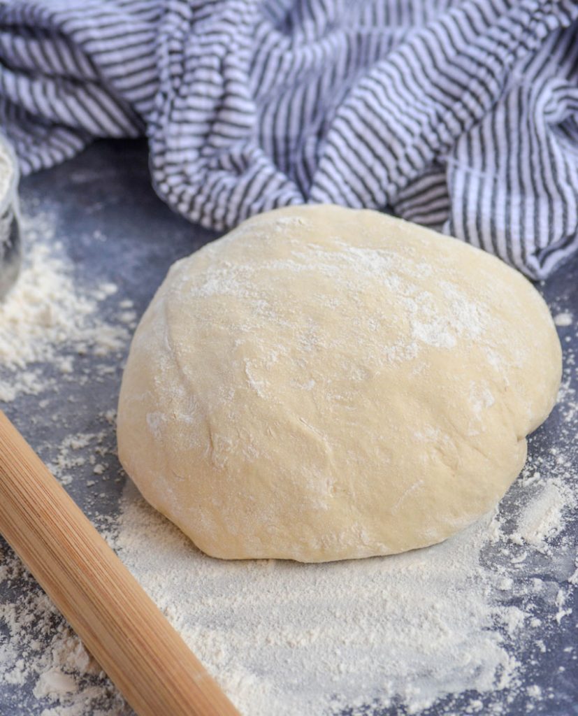 a ball of floured dough is shown on a gray back ground with a wooden rolling pin, excess flour, and a striped dish cloth in the back ground
