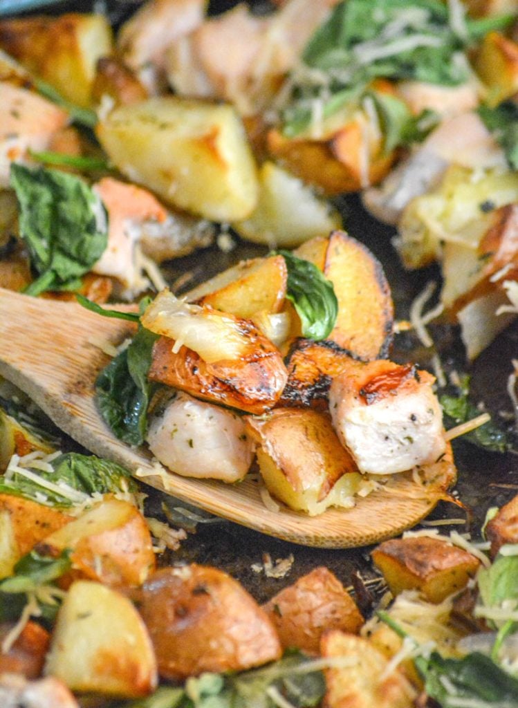 a wooden spoon with roasted potatoes, chicken chunks, white onion, and crispy spinach leaves on it