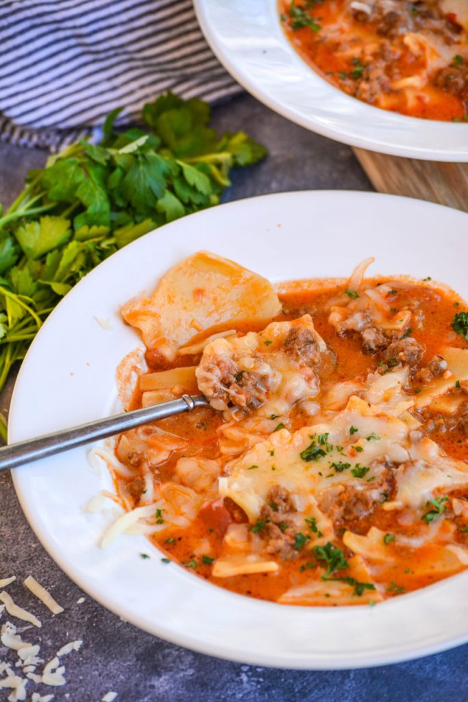 two bowls of lasagna soup, one with a spoon full, on a blue background with fresh parsley pictured