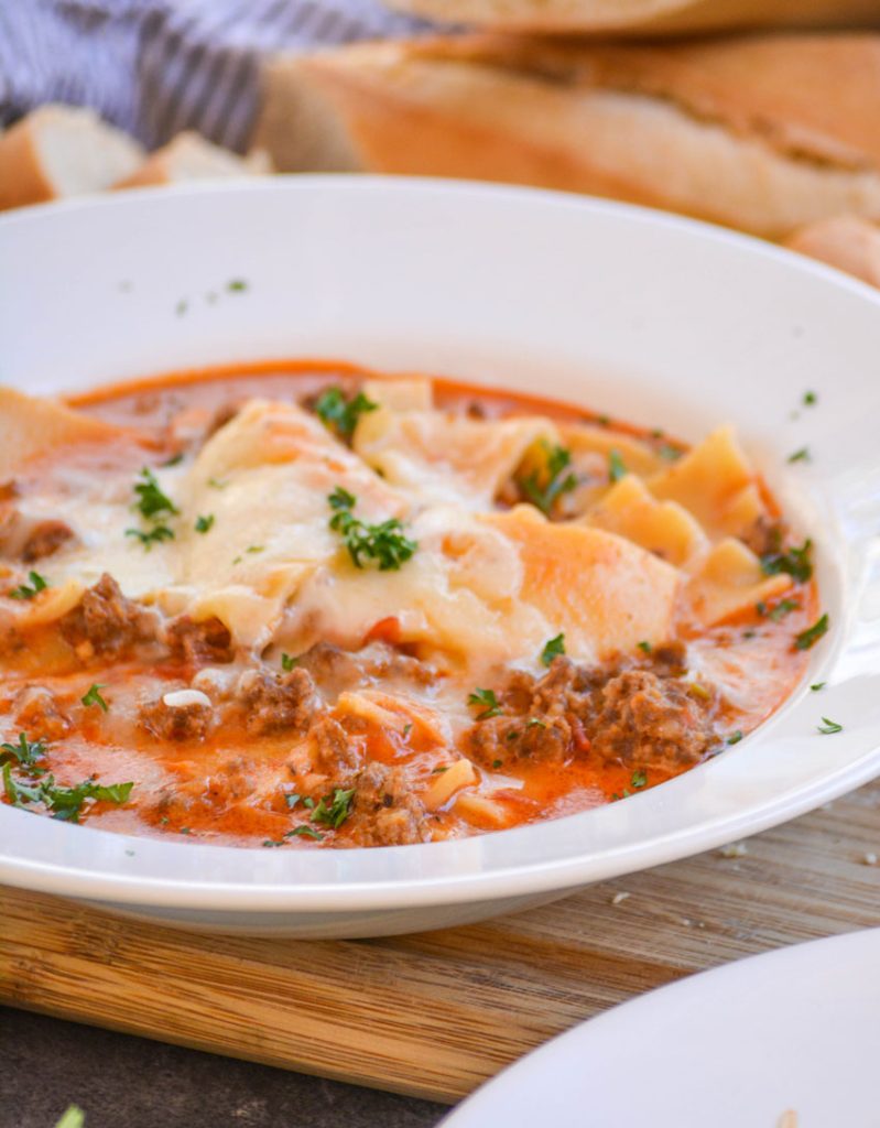 a white bowl full of lasagna soup with melted cheese on top and sprinkled with fresh parsley leaves