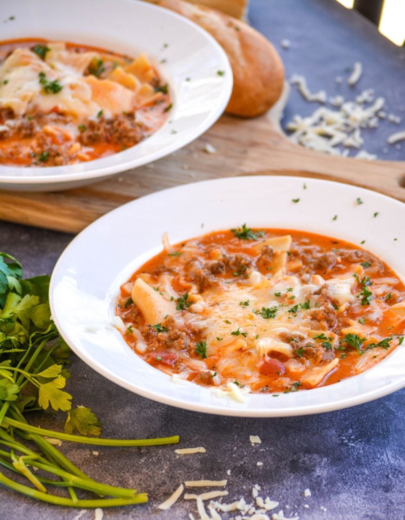 two bowls of lasagna soup on a blue background, the second on a cutting board, both pictured with melted cheese and parsley