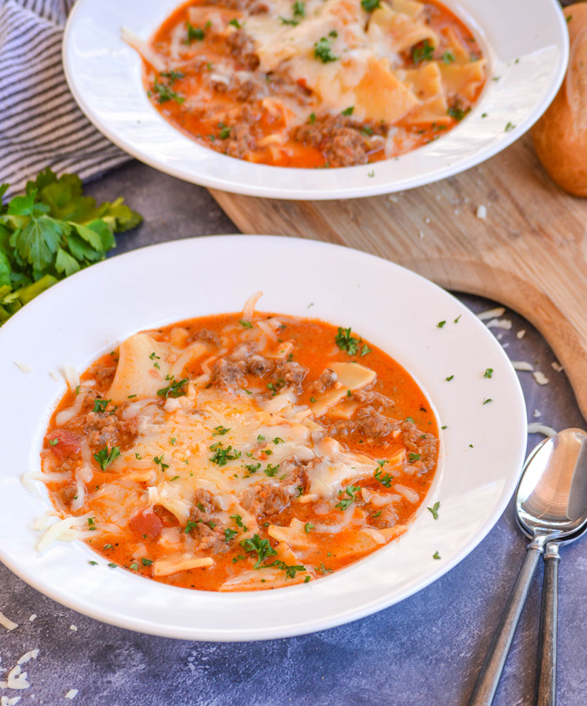 two bowls of lasagna soup on a blue background, the second on a cutting board with spoons pictured, both pictured with melted cheese and parsley sprinkled on top