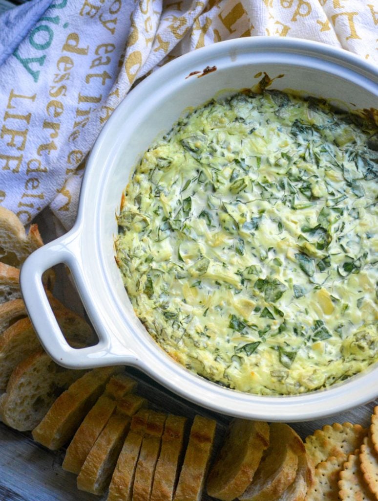 creamy spinach and artichoke dip in a white bowl surrounded by thinly sliced baguette bread