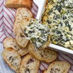 creamy spinach and artichoke dip on a slice of toasted baguette bread resting on the edge of a white baking dish
