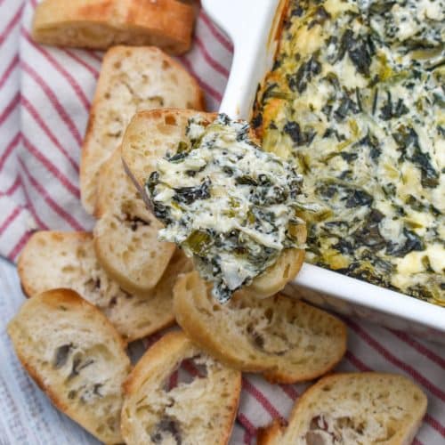 creamy spinach and artichoke dip on a slice of toasted baguette bread resting on the edge of a white baking dish