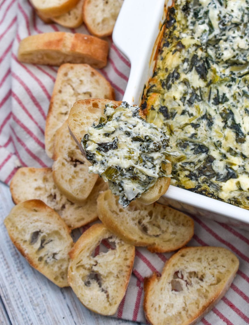 creamy spinach and artichoke dip on a slice of toasted baguette bread resting on the edge of a white baking dish