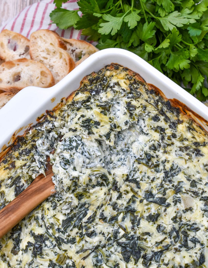 a wooden spoon scooping hot spinach and artichoke dip out of a white baking dish
