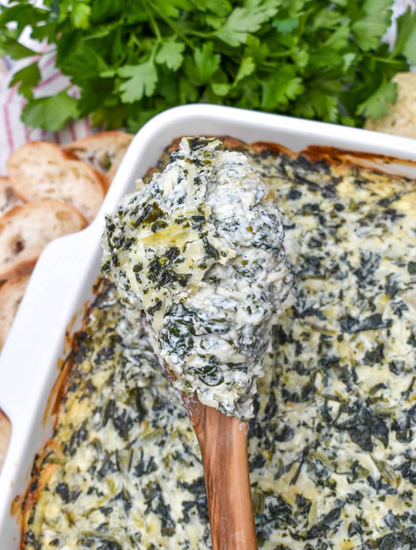 a wooden spoon holding up a scoop of hot creamy spinach and artichoke dip