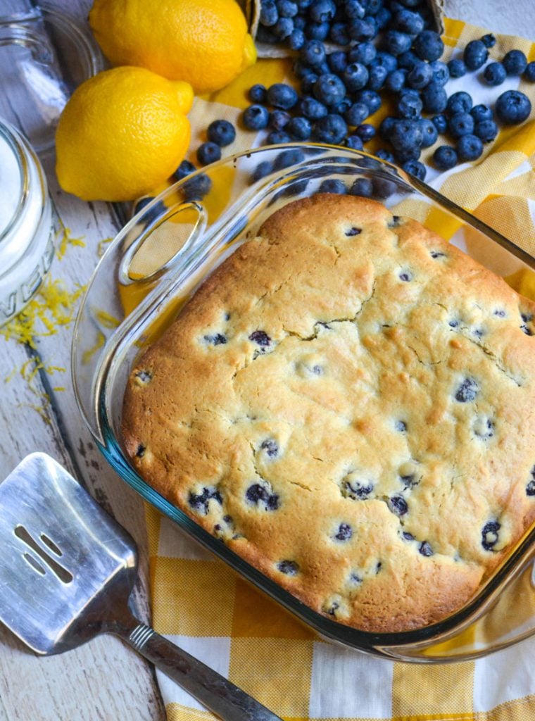 blueberry cake for breakfast on a yellow checkered napkin with fresh lemon, blueberries, and a metal spatula