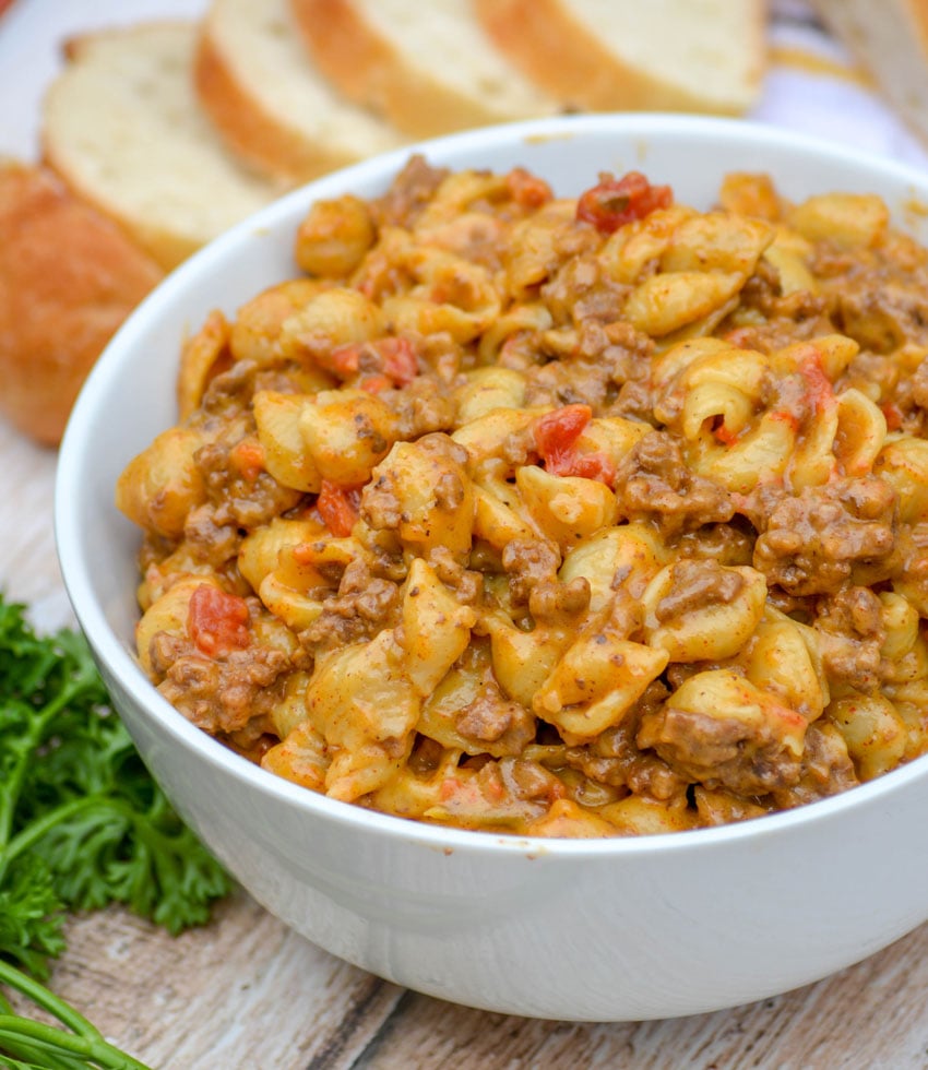 creamy cheesy shell pasta with ground beef and tomatoes in a white bowl on a wooden background with sliced bread and fresh parsley in the background