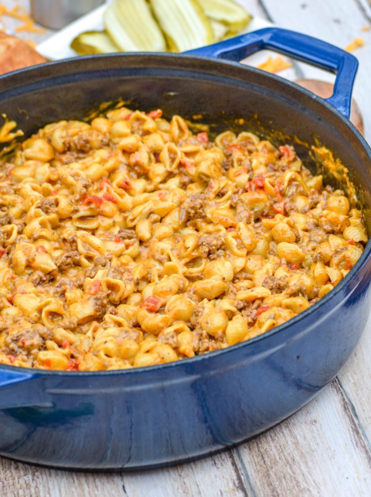 beef and shell shaped pasta with tomatoes in a cheesy sauce in a blue enameled dutch oven on a wooden background. Sliced dill pickles on a white plate & shredded cheddar cheese in the background.