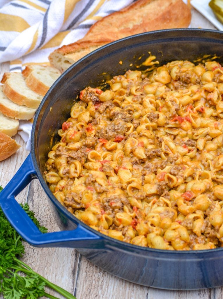 beef and shell shaped pasta with tomatoes in a cheesy sauce in a blue enameled dutch oven on a wooden background. Fresh parsley, a partially sliced baguette, and a striped dish cloth are in the background.