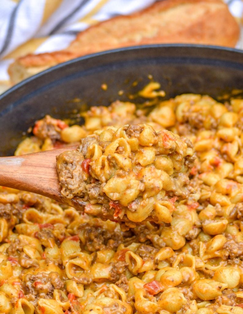 beef and shell shaped pasta with tomatoes in a cheesy sauce in a blue enameled dutch oven on a wooden background. A wooden spoon holds a generous scoop of macaroni with a partially sliced baguette, and a striped dish cloth are in the background.