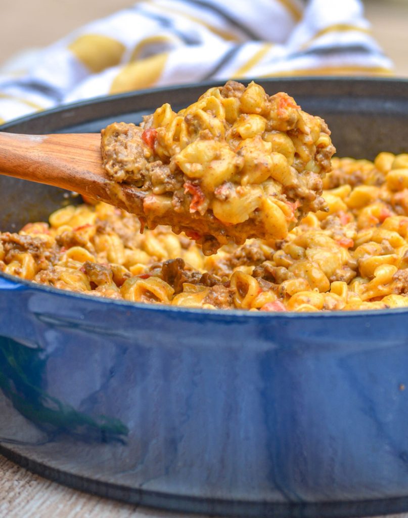 beef and shell shaped pasta with tomatoes in a cheesy sauce in a blue enameled dutch oven on a wooden background. A wooden spoon is shown holding a loft a hearty scoop of the pasta with a striped dish towel in the background.