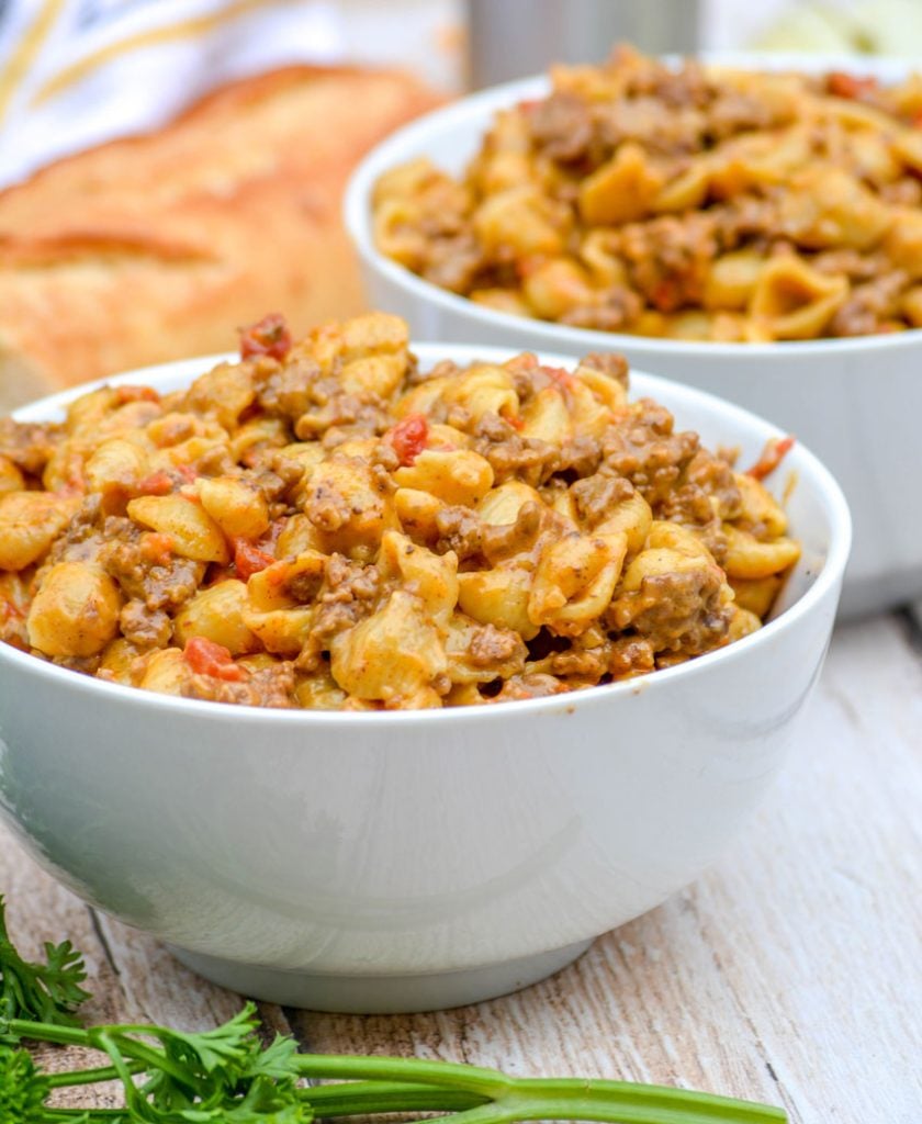 shell pasta in a creamy cheese sauce with ground beef and tomatoes is show in two white bowls on a wooden background with fresh parsley & bread in the background