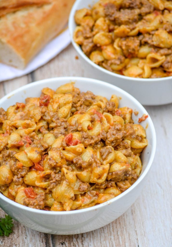 beef and shell shaped pasta with tomatoes in a cheesy sauce in two white bowls on a wooden background with a fresh baguette in the background.