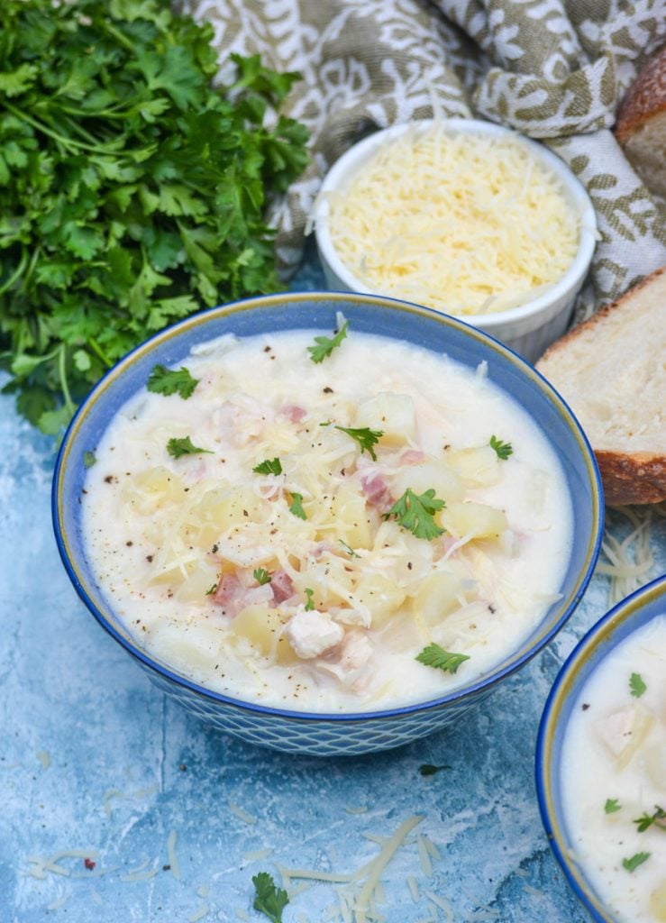 soup in a blue bowl on a blue backdrop with fresh parsley, shredded cheese, and bread in the background