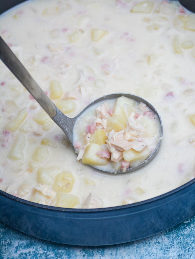 a silver ladle holding a scoop full of chicken cordon bleu soup with chicken, potatoes, and ham all showing