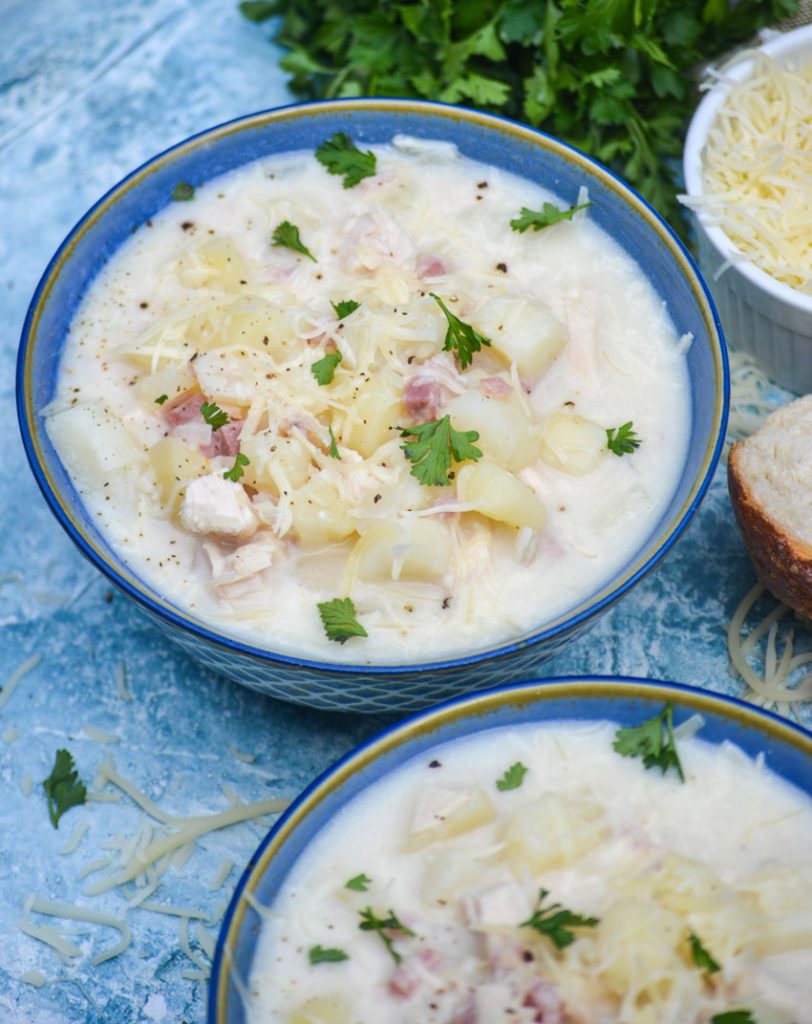 chicken cordon bleu soup in two blue bowls on a blue background with fresh parsley and shredded Swiss cheese off to the side