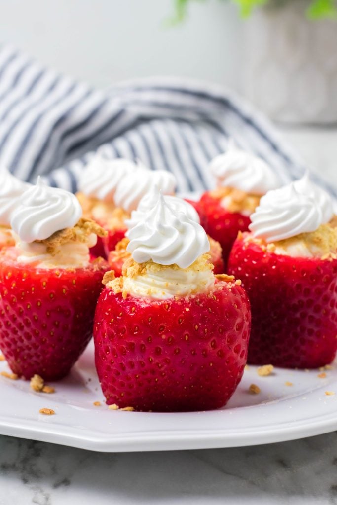 whipped cream topped cheesecake stuffed strawberries on a white plate on marble counter with striped dish cloth and plant in background