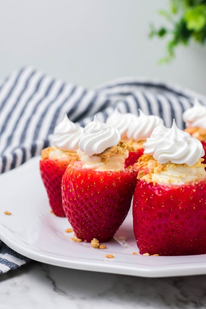 cheesecake stuffed strawberries on a white plate on marble background with a striped dish cloth and plant behind them