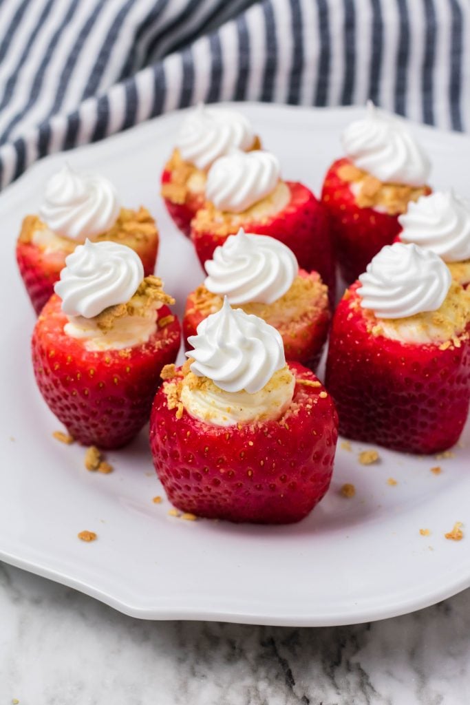 cheesecake stuffed strawberries on a white place with graham cracker crumbs on marble background with striped dish towel