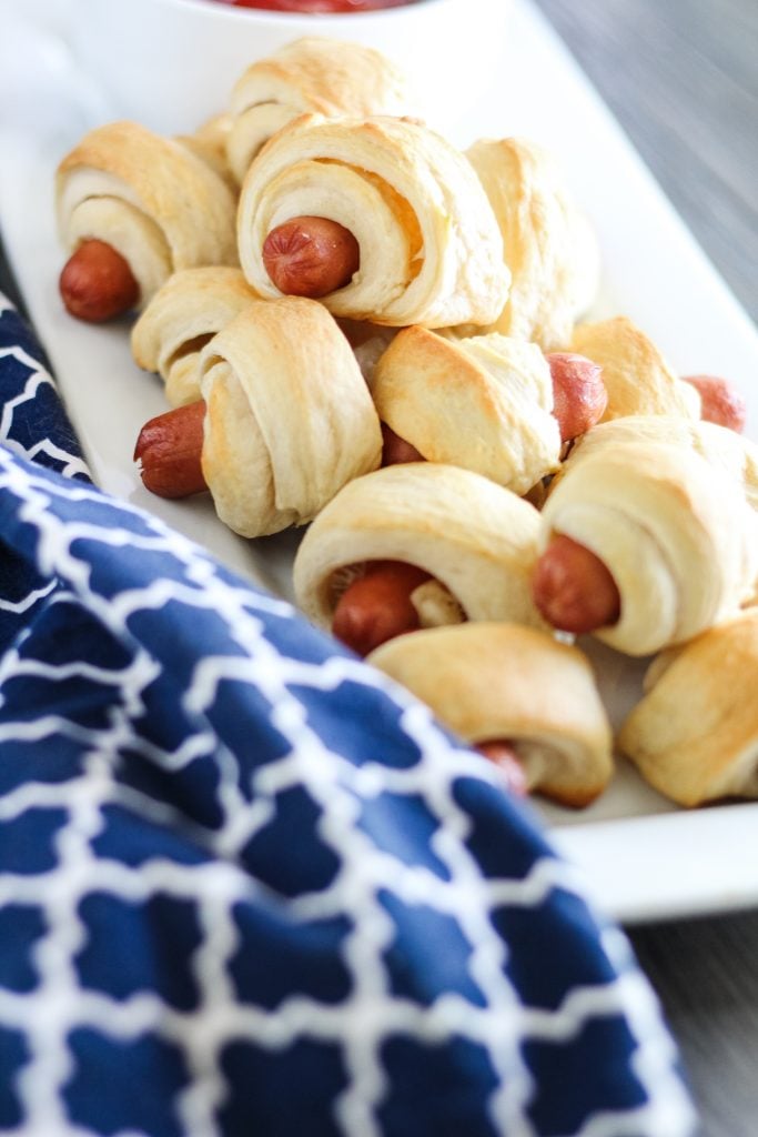 classic pigs in a blanket piled high on a white rectangular serving platter with a bowl of ketchup for dipping