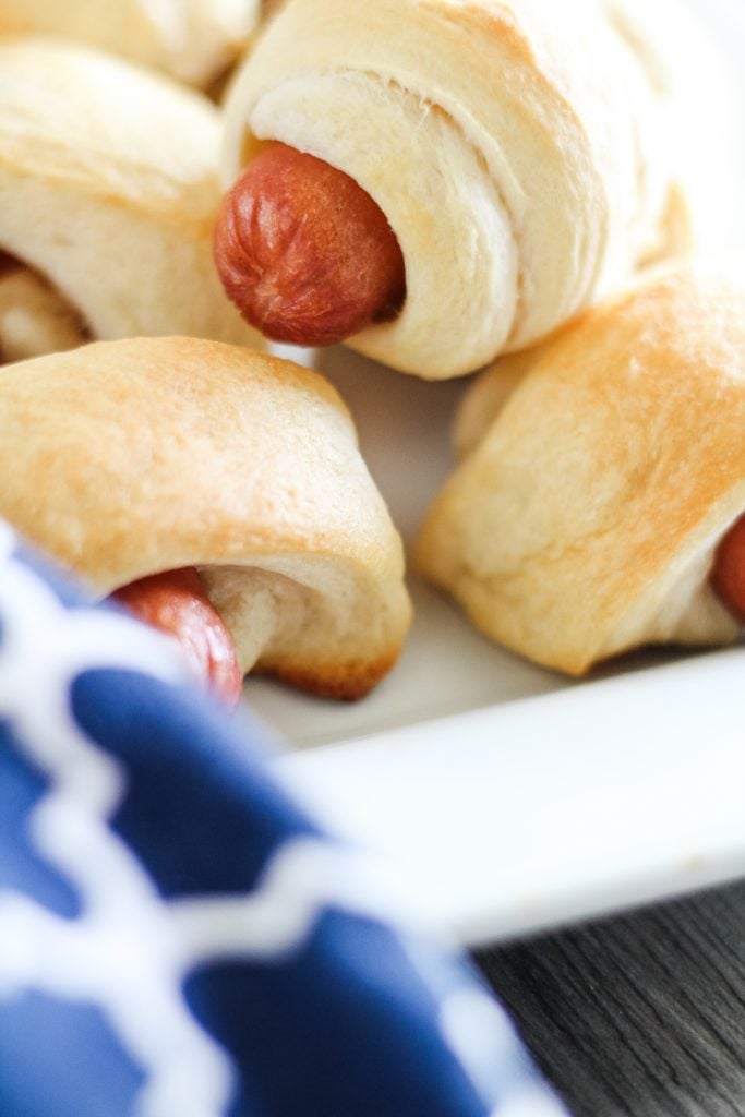 classic pigs in a blanket piled high on a white rectangular serving platter with a bowl of ketchup for dipping