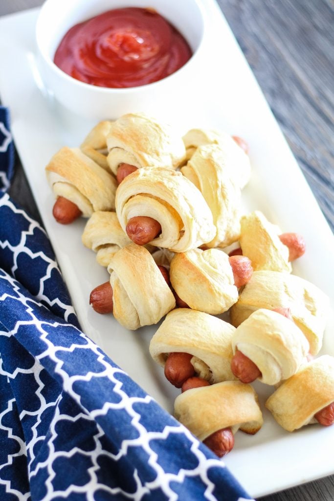 classic pigs in a blanket piled high on a white rectangular serving platter with a bowl of ketchup for dipping