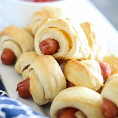 classic pigs in a blanket piled high on a white rectangular serving platter with a bowl of ketchup for dipping