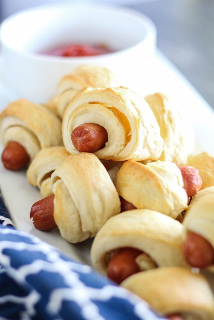 classic pigs in a blanket piled high on a white rectangular serving platter with a bowl of ketchup for dipping