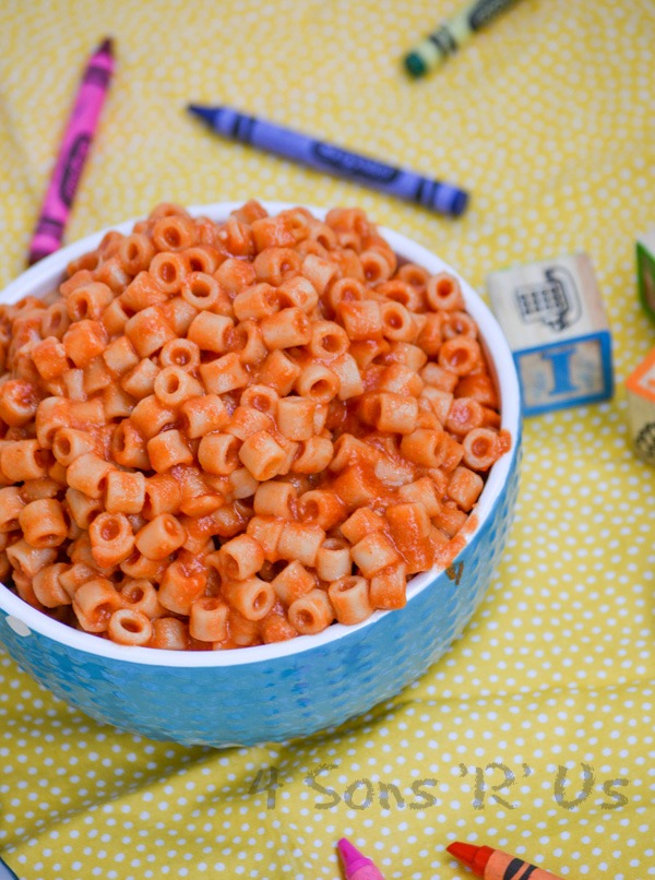 Homemade Spaghetti O's in a white rimmed blue bowl on a yellow background with crayons and wooden toy blocks