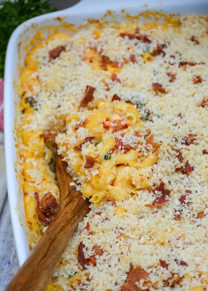 a wooden spoon digging into a blt flavored macaroni and cheese casserole in a white baking dish