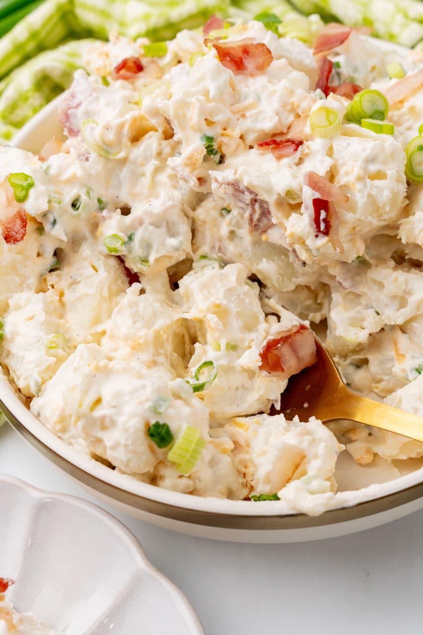 a golden spoon scooping loaded potato salad out of a glass mixing bowl