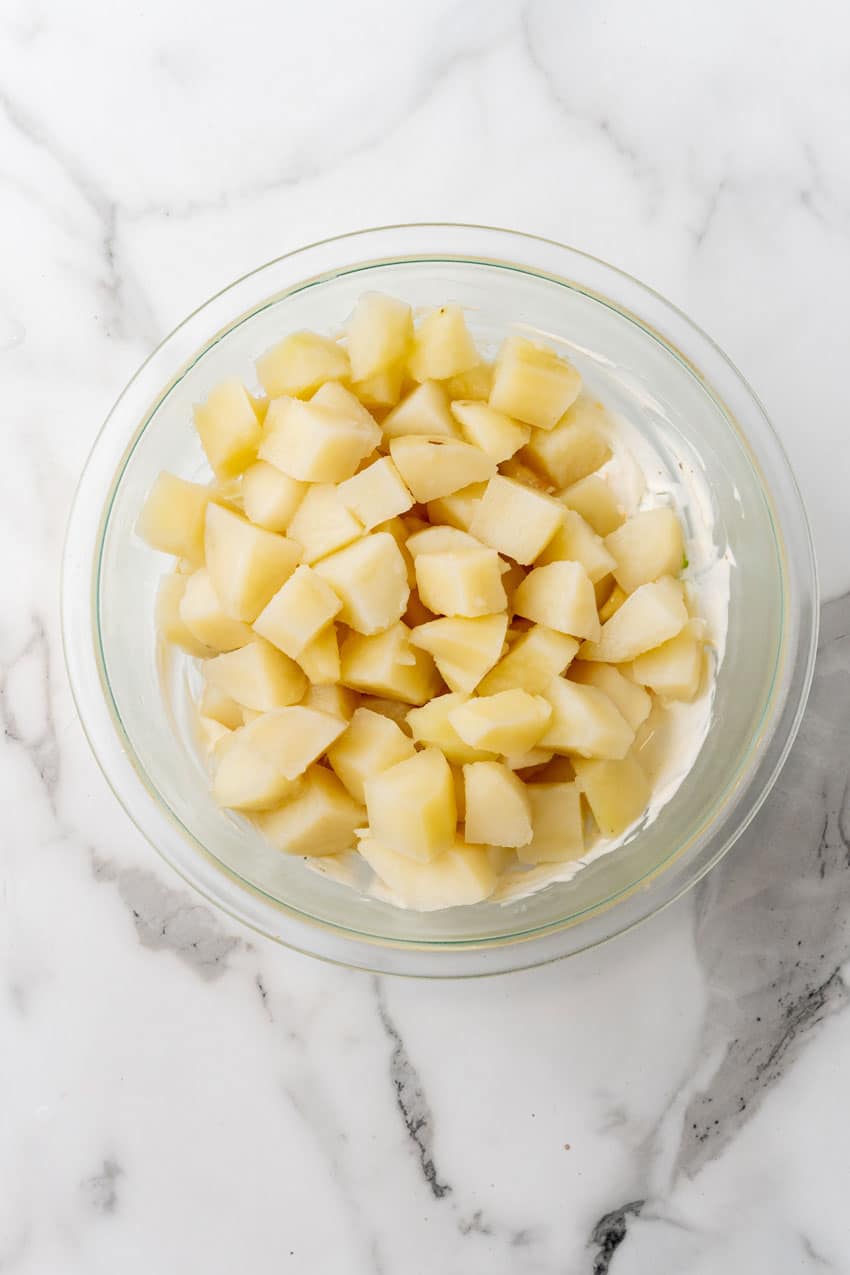 diced cooked chunks of potatoes sitting on potato salad dressing in a glass mixing bowl