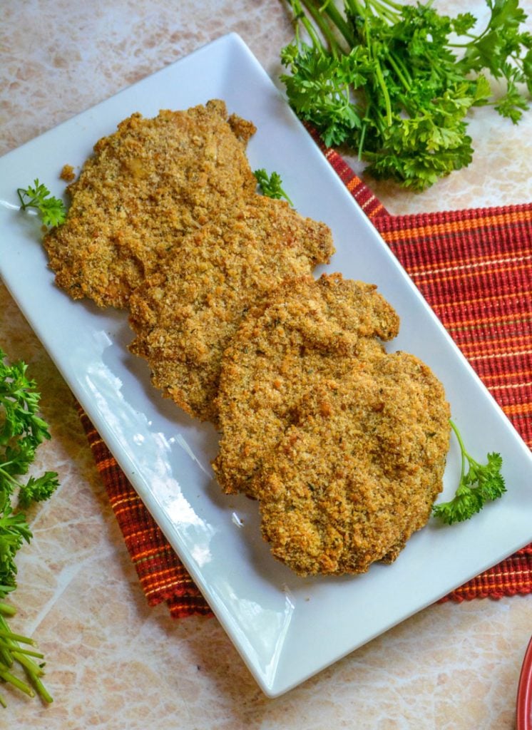 four Crispy, Baked Italian Ranch Pork Chops shown on a rectangular white serving dish on a red striped napkin with bunches of fresh parsley in the background