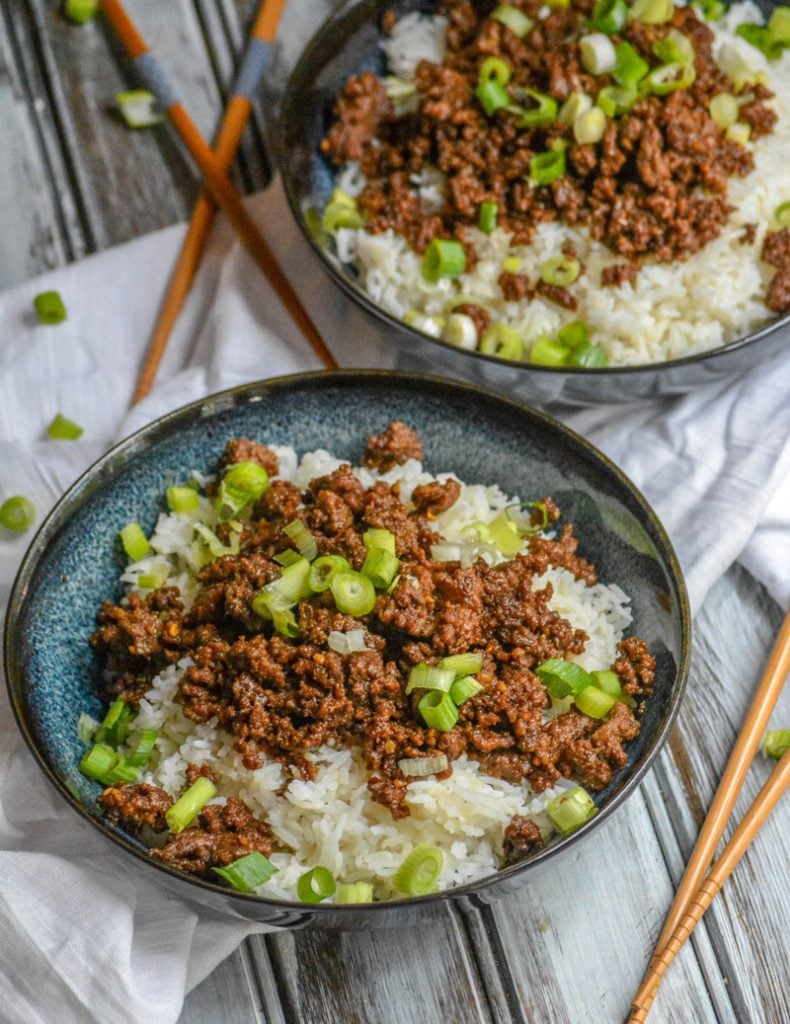 Korean Barbecued Beef with sliced green onions over steamed white rice in blue bowls with sets of chop sticks in the background