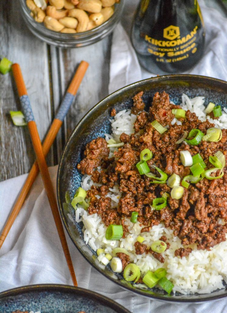 Korean Beef on a bed of white rice with chop sticks, soy sauce, and nuts in the background