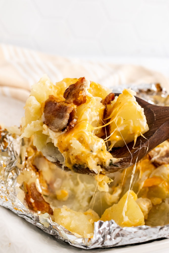 a wooden spoon shown lifting a scoop of three cheese potatoes from a grilled foil packet