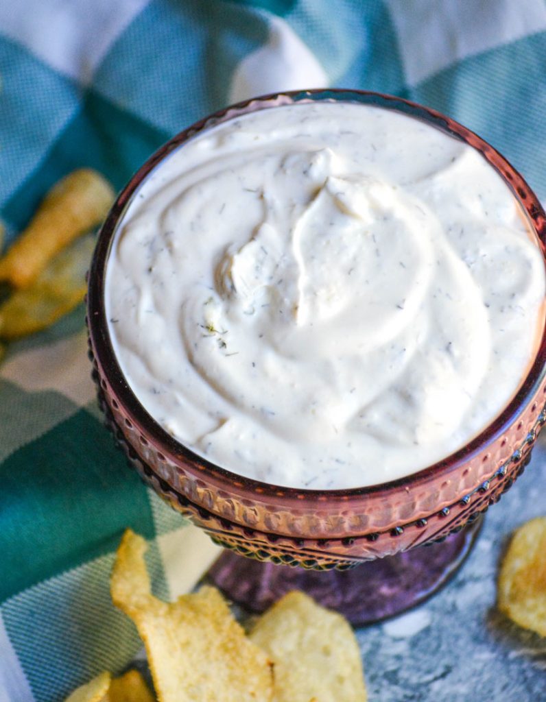 dill dip in a purple glass serving dish with a teal checkered cloth napkin and crisp chips in the background