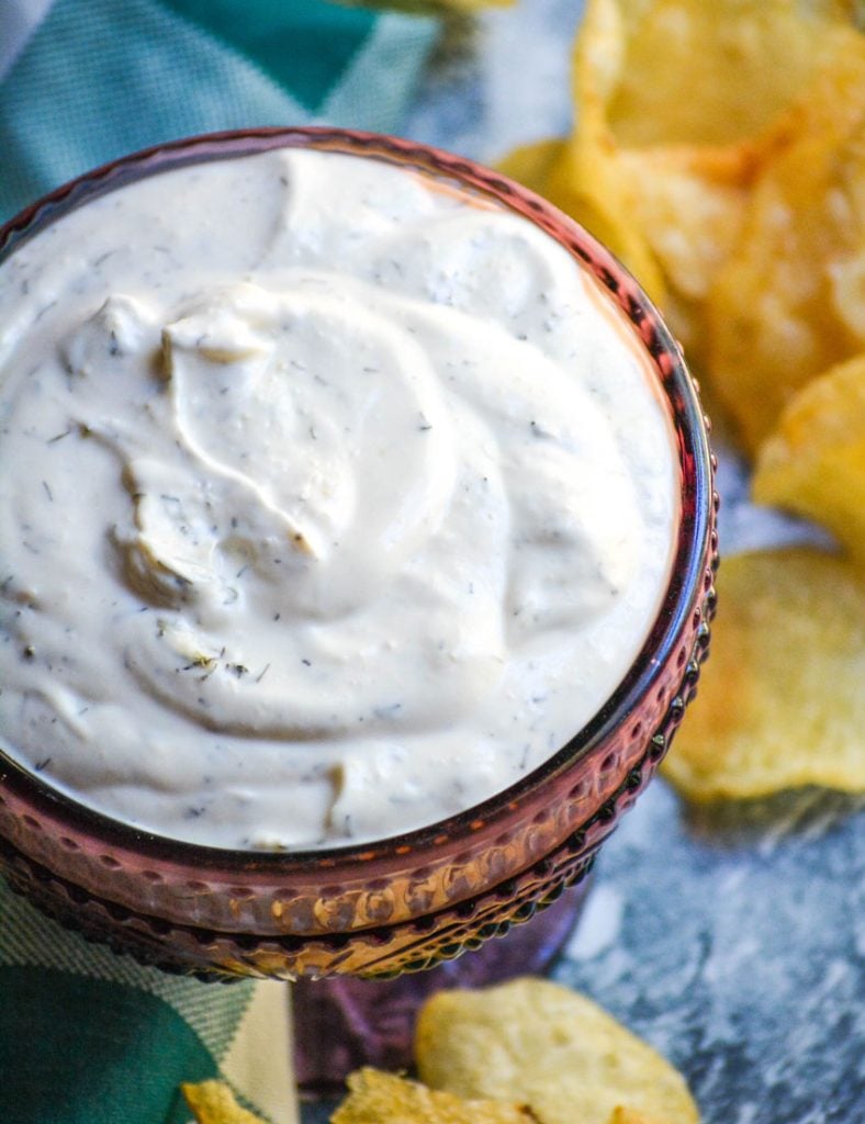 dill dip in a purple glass serving dish with a teal checkered cloth napkin and crisp chips in the background