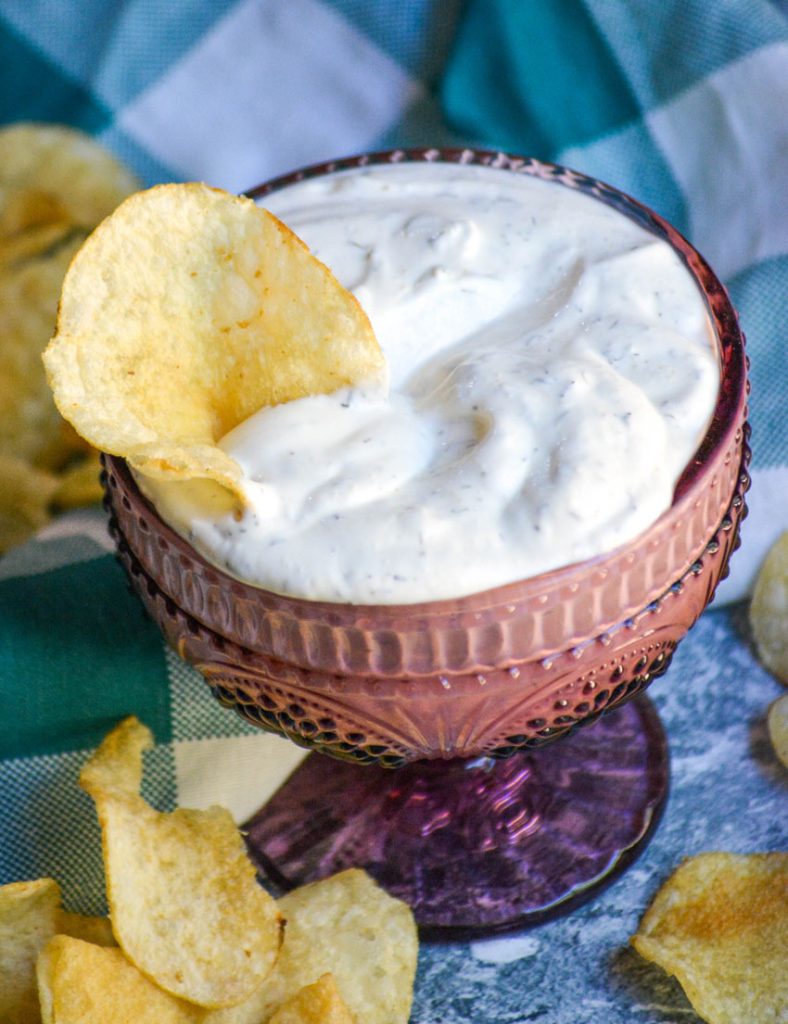 creamy dill dip in a purple glass serving dish with a potato chip stuck in for dipping