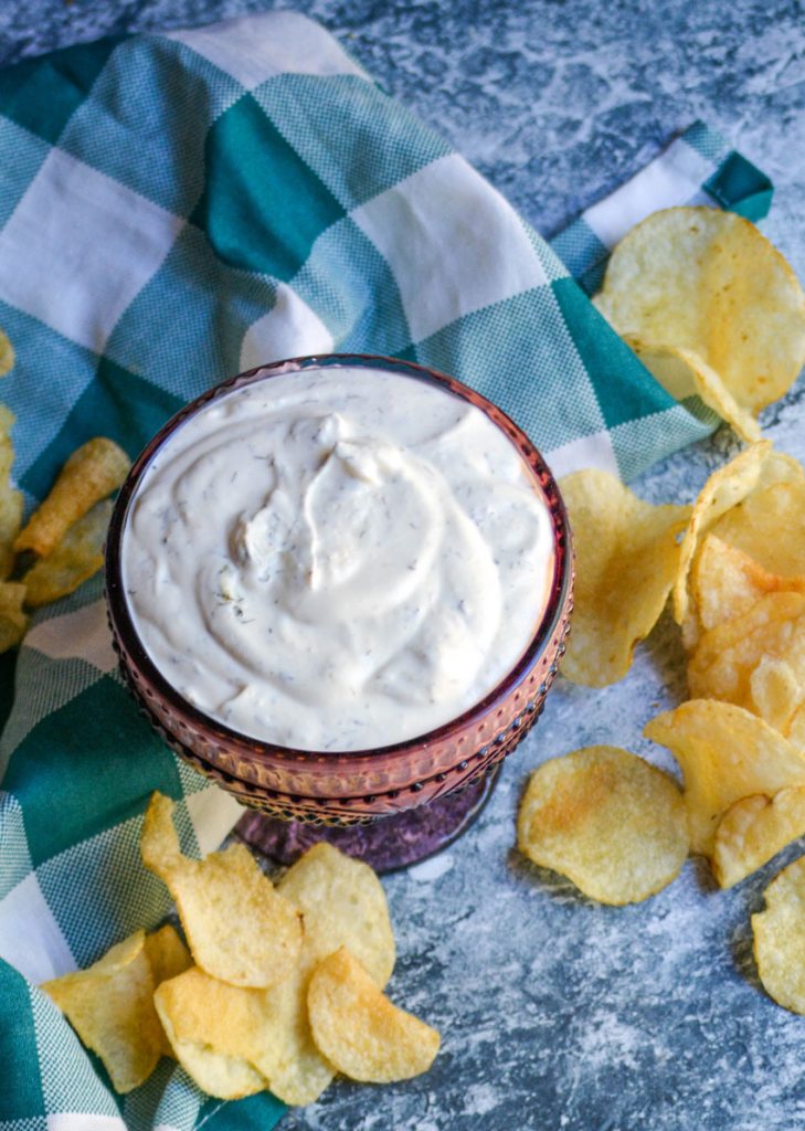 dill dip in a purple glass serving dish with a teal checkered cloth napkin and crisp chips in the background