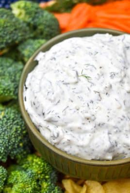 creamy dill dip recipe in a small brown bowl surrounded by an assortment of fresh vegetables and crispy potato chips
