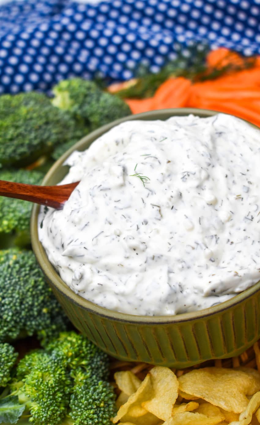 a wooden spoon scooping creamy dill dip out of a small brown bowl surrounded by fresh vegetables