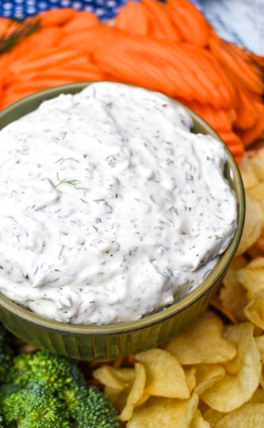 creamy dill dip recipe in a small brown bowl surrounded by an assortment of fresh vegetables and crispy potato chips