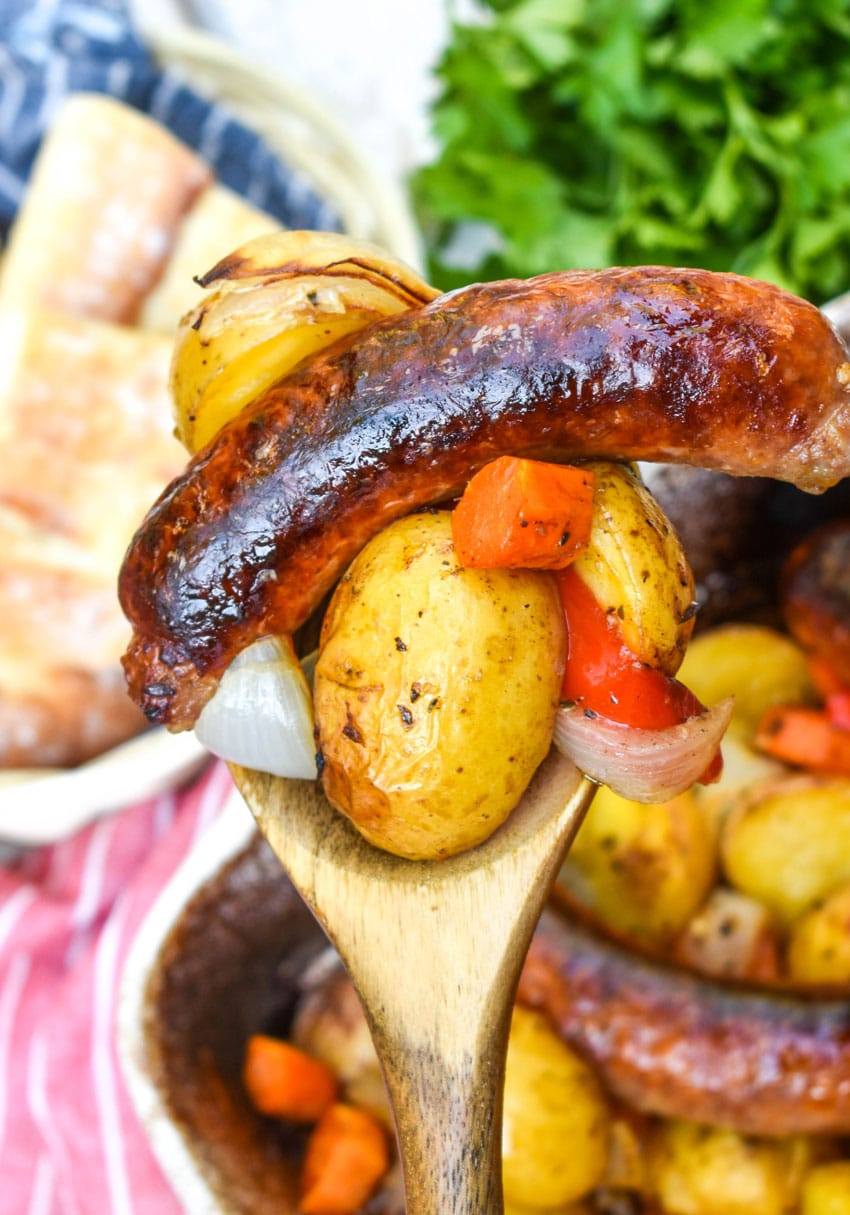 a wooden spoon holding up a scoop of baked Italian sausage and vegetables